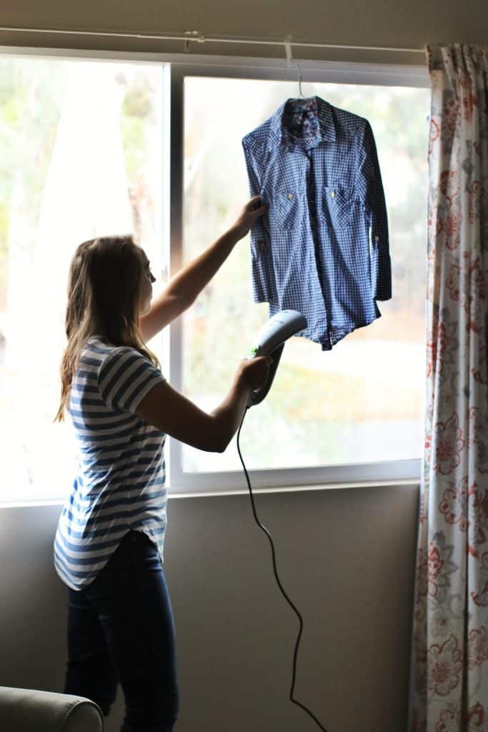 A woman uses a handheld steamer to steam a shirt hanging in the window